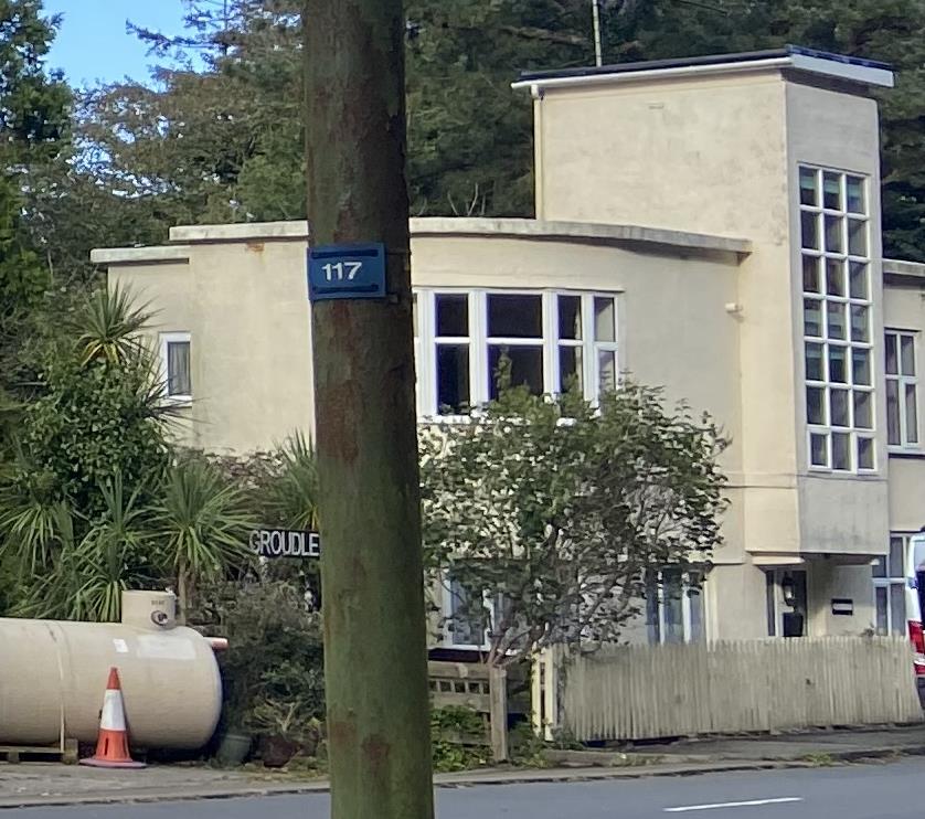 A street-level photograph showing a cream-colored, modernist-style detached house with large windows and a flat roof. A wooden utility pole with house number 117 is prominent in the foreground.