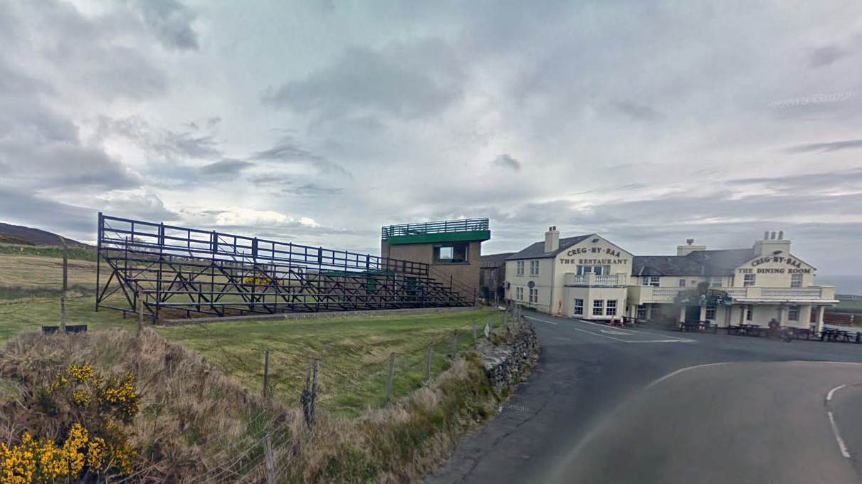 A photograph showing a large metal grandstand structure situated next to a road, with a building labeled 'Creg-ny-Baa' in the background.