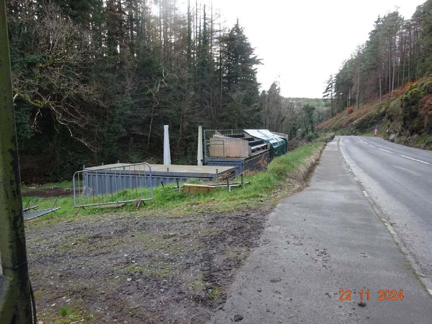 A photograph showing a roadside area with temporary structures, including a raised platform with railings and container-like units, set against a wooded background.