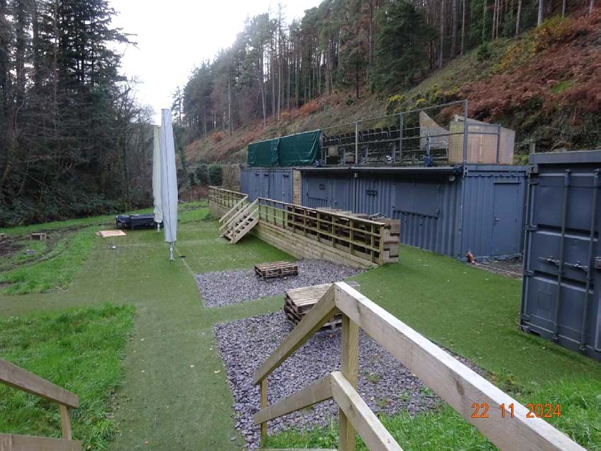 A photograph showing a row of grey shipping containers built into a hillside with wooden decking, retaining walls, and artificial turf.