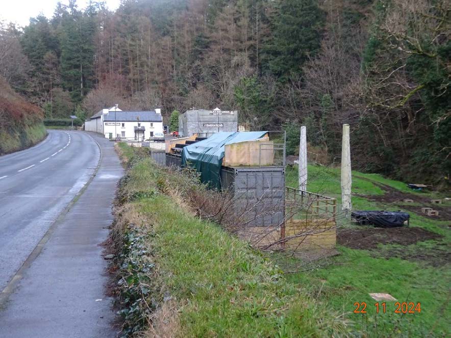A roadside photograph showing a row of shipping containers and wooden posts next to a paved path, with a white building and forest in the background.