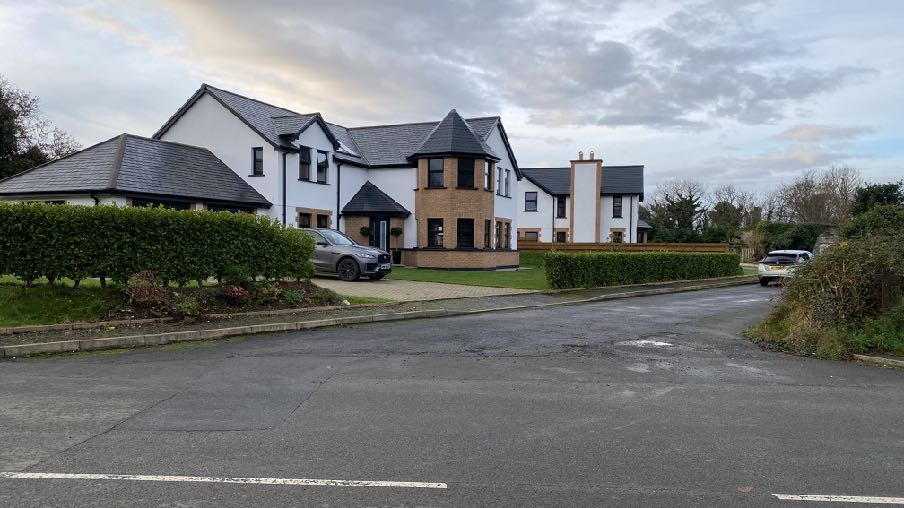 A large, modern detached house with white render and a slate roof is viewed from the street, featuring a paved driveway and green hedges.