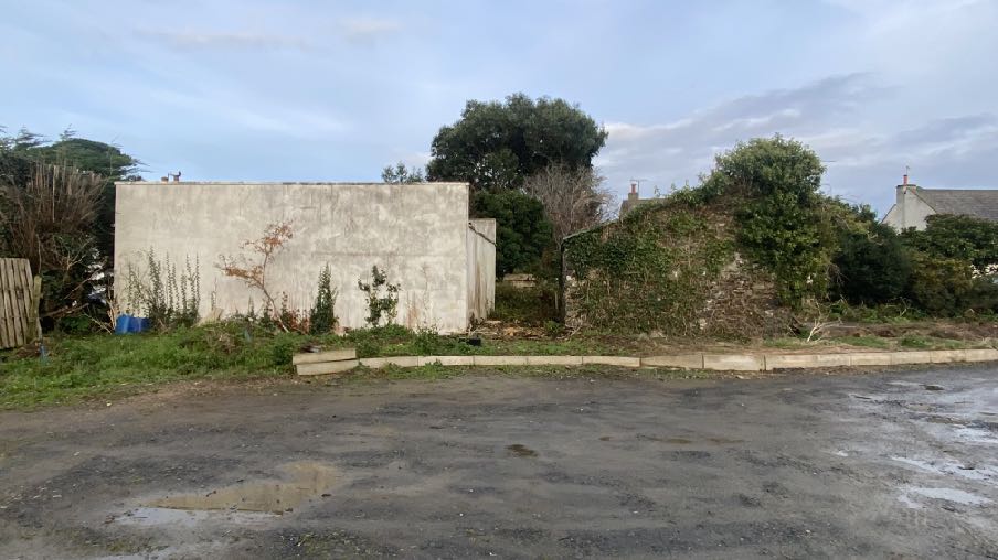 A photograph of a rural site showing a paved foreground area, likely an access road, with a large plain wall and an ivy-covered stone wall in the background.