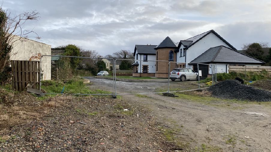 A photograph showing a modern detached house behind temporary metal fencing, with a gravel driveway and construction materials in the foreground.