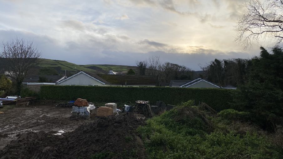 A photograph of a muddy construction plot with building materials, bordered by a hedge and existing houses against a rural hillside backdrop.