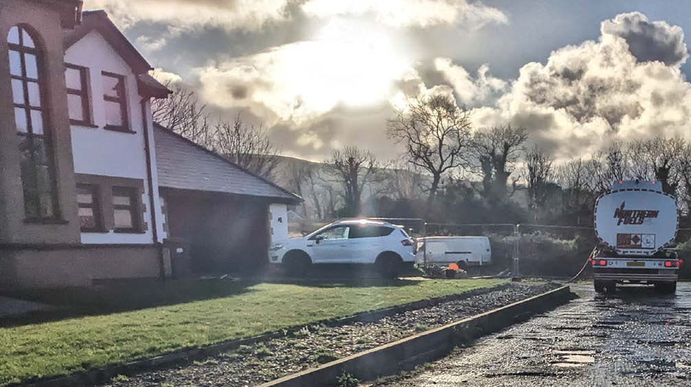 A photograph showing a white detached house with an attached garage on the left, a white SUV parked on the grass, and a fuel tanker truck on a wet road in a rural setting.
