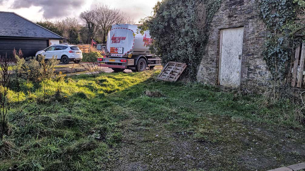 A photograph showing a rural site with a grassy foreground, a large white agricultural tanker truck, a parked white car, and stone buildings covered in ivy.