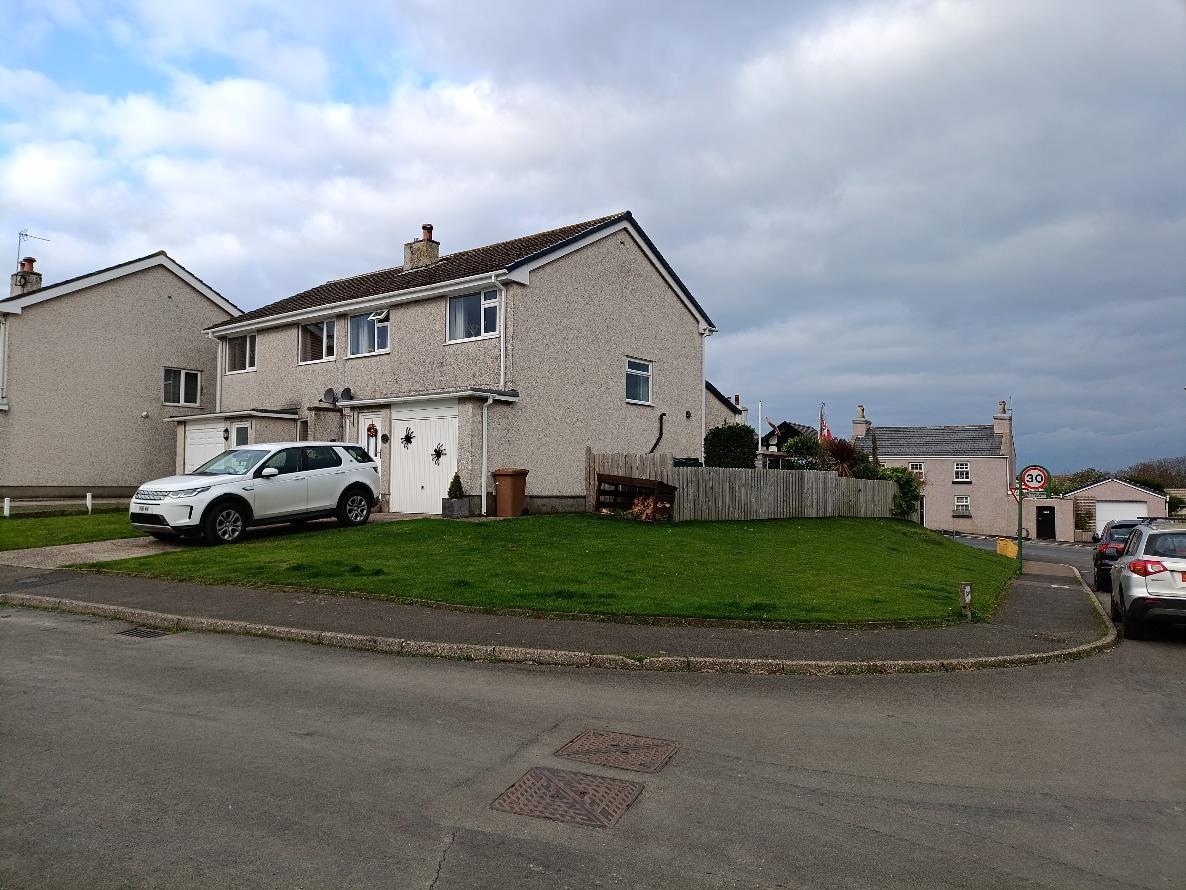 A street-level photograph showing a two-story detached house with a driveway, a white car parked, and a wooden fence along the side boundary.