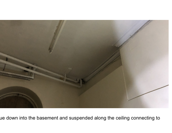 An interior photograph showing white pipes running along a ceiling and wall corner, likely illustrating the proposed internal downpipe installation.