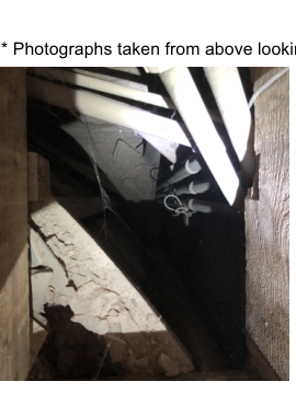A photograph taken from above looking into a roof void, showing wooden beams, insulation, and hanging cables or pipes.