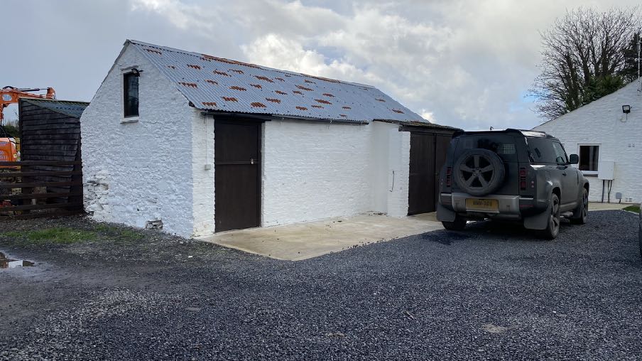 A photograph showing a white single-story outbuilding with a corrugated roof and a car parked on a gravel driveway, with construction machinery visible to the left.