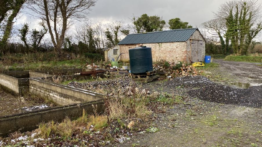 A photograph of a rural site featuring concrete drainage beds in the foreground, a large blue water tank, and a stone outbuilding with a dark roof in the background.