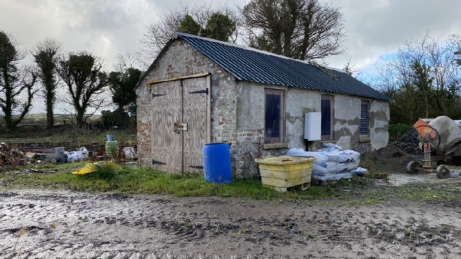 A photograph of a stone barn-like building with a slate roof, surrounded by construction materials and a cement mixer in a rural setting.