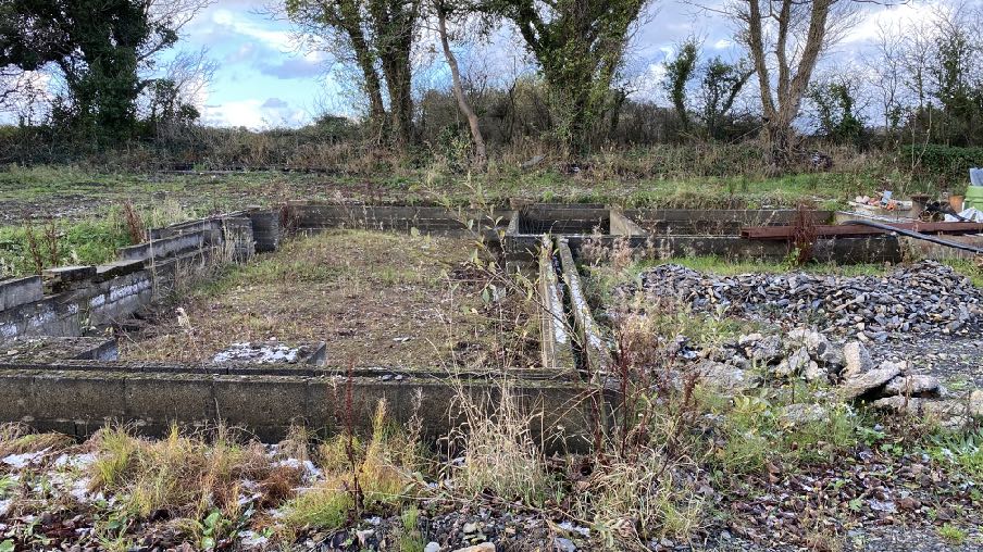 A photograph showing an overgrown concrete foundation base on a rural site, likely the existing ground conditions for the proposed stables.
