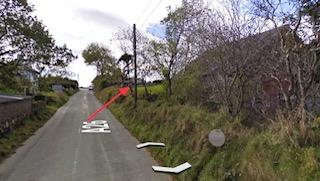 A photograph of a rural road with a red arrow pointing to a specific spot on the grassy verge, likely indicating the site for the proposed development.