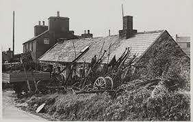 A black and white photograph showing existing rural farm buildings and agricultural equipment in the foreground.
