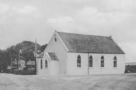 A black and white photograph showing a white, single-story building with a pitched roof and arched windows, resembling a small chapel or converted barn in a rural setting.