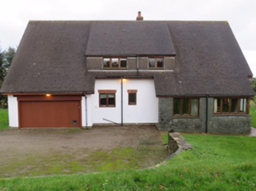 A photograph of a large, single-story rural building with a pitched roof, featuring a garage door, mixed stone and rendered walls, and a driveway.