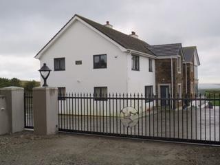 A photograph of a newly constructed white farmhouse with a brick extension, viewed from behind a black metal fence in a rural setting.