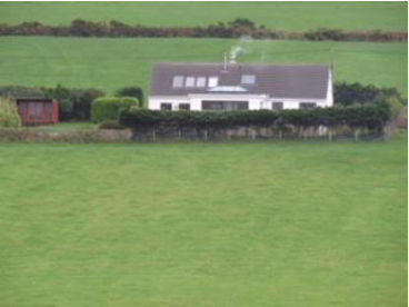 A distant photograph showing a white farmhouse with a dark roof situated in a green rural field, with a smaller outbuilding visible to the left.