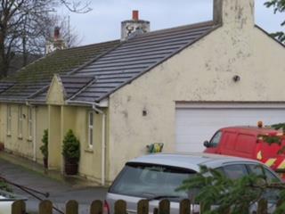 A photograph showing the exterior of an existing single-story building with a garage and parked vehicles, situated in a rural setting.