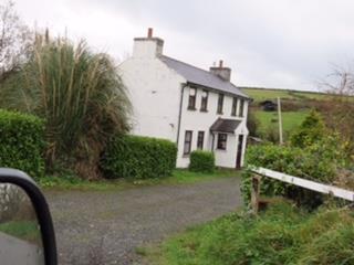 A photograph showing a white, two-story detached house with a slate roof and chimneys, situated on a gravel driveway in a rural setting with green fields in the background.