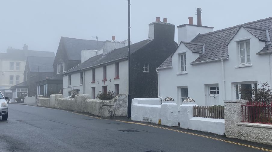 A misty street scene showing a row of traditional stone and whitewashed cottages with slate roofs and low stone boundary walls.