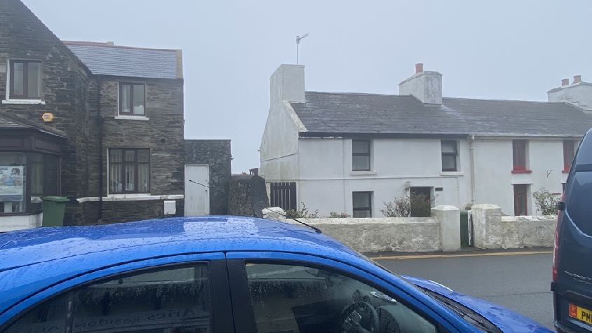 A street-level photograph showing existing stone and white-washed buildings in a village setting, with a blue car in the foreground.