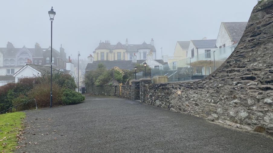 A photograph showing a paved road or driveway leading uphill past a high stone wall, with residential houses visible in the misty background.
