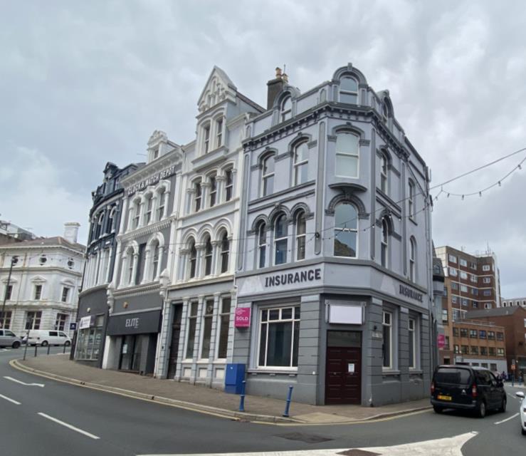 A street-level photograph of a grey corner commercial building with 'INSURANCE' signage, situated next to white and black buildings on a curved street.