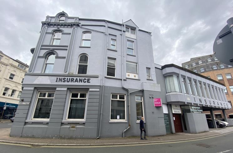 A street-level photograph of a grey, multi-story commercial building with 'INSURANCE' signage on the facade, situated on a street corner in an urban setting.
