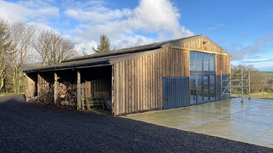 A photograph of a wooden agricultural building undergoing conversion, featuring new large windows, scaffolding, and a concrete patio area in a rural setting.