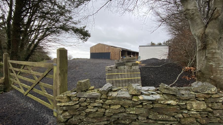 A photograph of a rural farm entrance featuring a wooden gate and stone wall. In the background, a large wooden agricultural building stands on a gravel driveway.