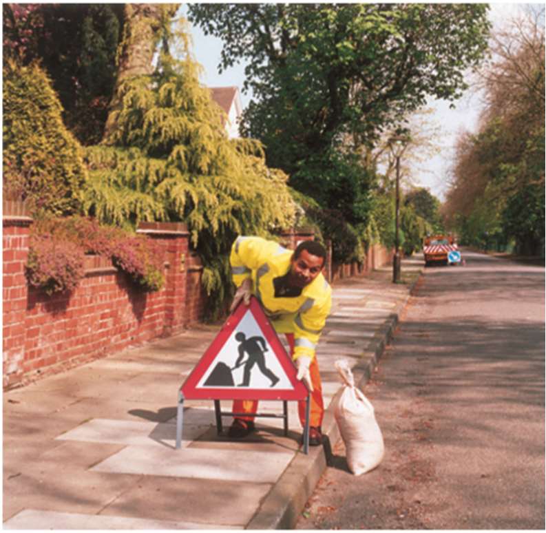 A stock photograph showing a road worker in high-visibility clothing placing a triangular road works sign on a residential pavement next to a brick wall.