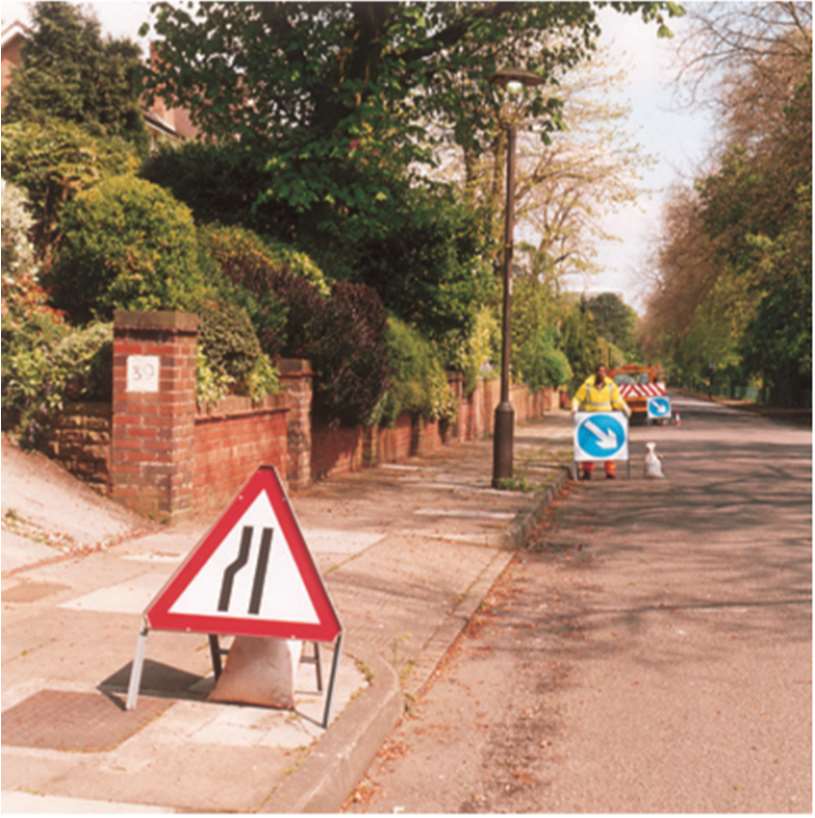 A photograph of a residential street featuring road work signage, including a narrowing road warning sign in the foreground and a traffic marshal holding a keep left sign further down the road.