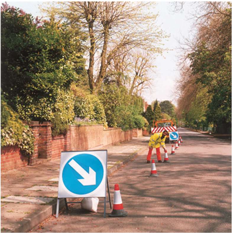 A photograph showing roadworks on a residential street with traffic cones, directional signs, and a worker in high-visibility clothing.