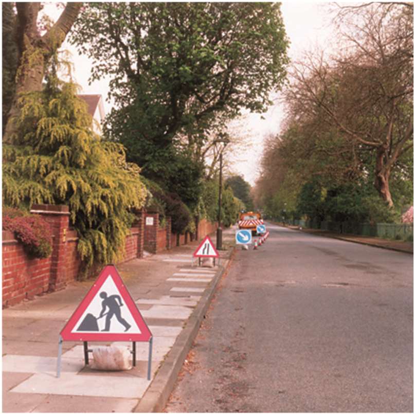 A photograph showing a residential street with temporary road work signs and traffic barriers set up on the pavement and road.