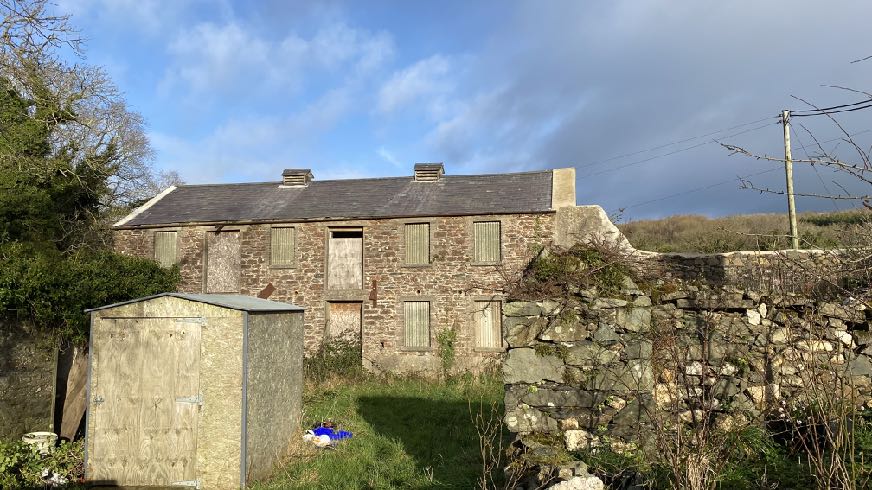A photograph showing a dilapidated stone agricultural building with boarded windows and doors, situated next to a wooden shed and stone wall in a rural setting.