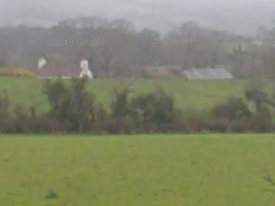 A low-resolution photograph showing a rural green field in the foreground with trees and a white building or agricultural structure visible in the distance.