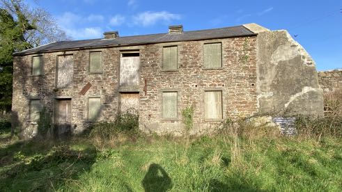 A photograph of a long, two-story stone building in a state of disrepair with boarded-up windows and doors, surrounded by overgrown vegetation.