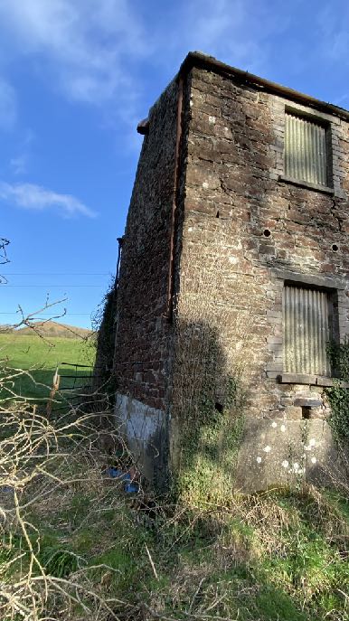 A photograph showing a dilapidated stone building, likely an old barn, with corrugated windows and overgrown vegetation in a rural setting.