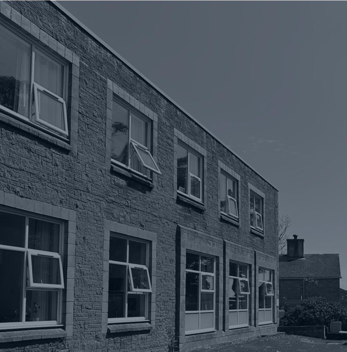 A monochromatic photograph showing the exterior facade of a two-story stone building with multiple windows, likely the Buchan School mentioned in the application.