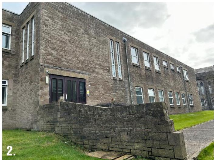 Exterior photograph of a large stone building, likely a school, featuring a dark entrance door and a low stone retaining wall in the foreground.