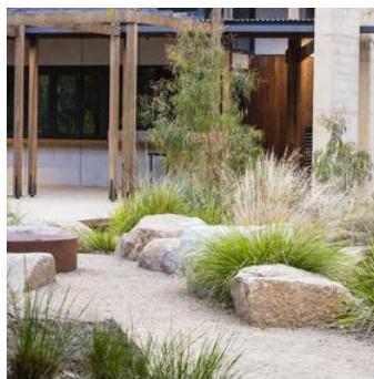A photograph showing a landscaped courtyard area featuring large rocks, ornamental grasses, and a gravel path. A modern building structure with wooden posts and large windows is visible in the background.