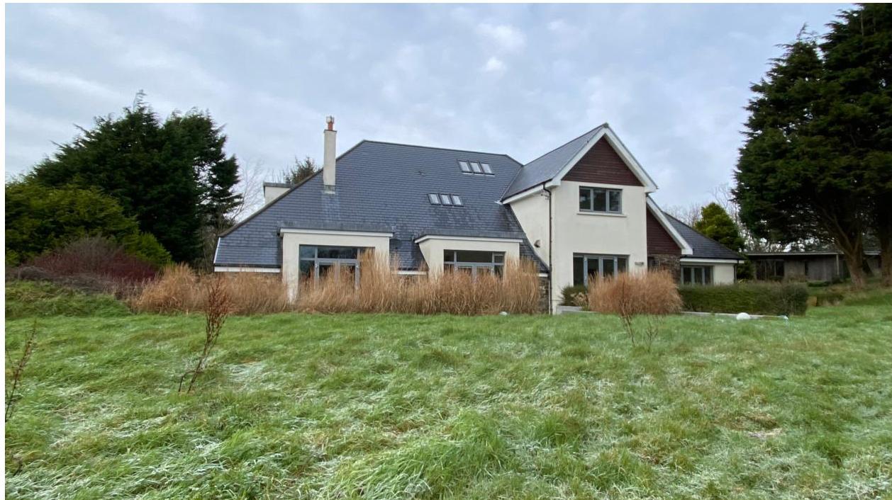 A photograph showing the exterior of a large detached house with a slate roof, situated in a grassy, rural setting with mature trees.