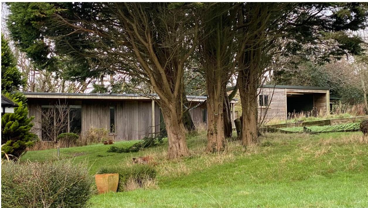 A photograph showing a single-story wooden building, likely a bungalow, partially hidden by large trees. To the right, a separate wooden outbuilding or garage stands in a grassy, rural setting.