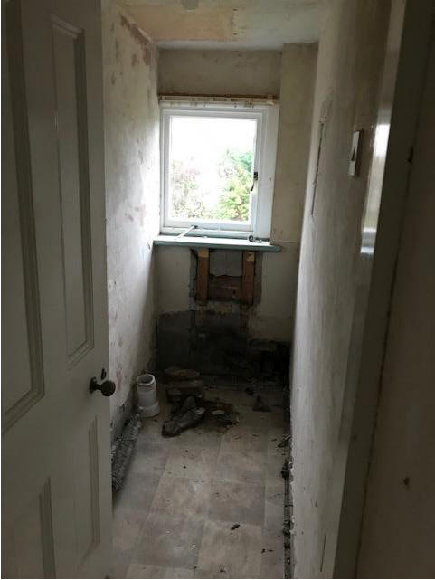 Interior photograph of a room undergoing demolition or renovation, showing stripped walls, debris on the floor, and a window at the far end.