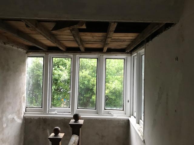 Interior photograph of a room under construction with exposed wooden ceiling joists and a row of white windows overlooking greenery.