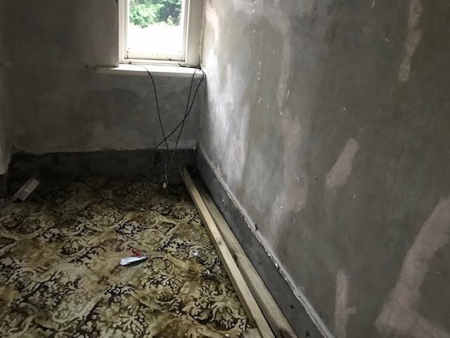 Interior view of a room with unfinished walls and patterned carpet, likely showing the existing property condition.