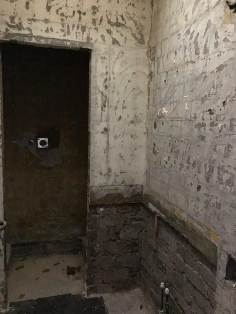 Interior photograph of a room in an existing house showing exposed brickwork on the lower walls and peeling plaster above, likely documenting the property to be demolished.